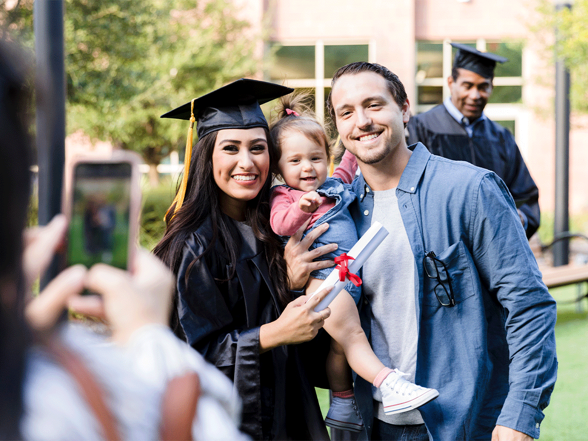 Graduating student in her cap and gown, taking a picture with her husband and baby.