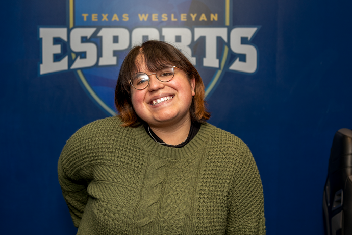 Texas Wesleyan student Cindy Flores wearing a green sweater posing in front of a logo that says texas wesleyan esports