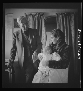 Woman sitting in a room with a baby in her lap. Older man is standing holding the baby's hand and looking at the baby. Baby is looking at the man. Woman is looking at the baby.