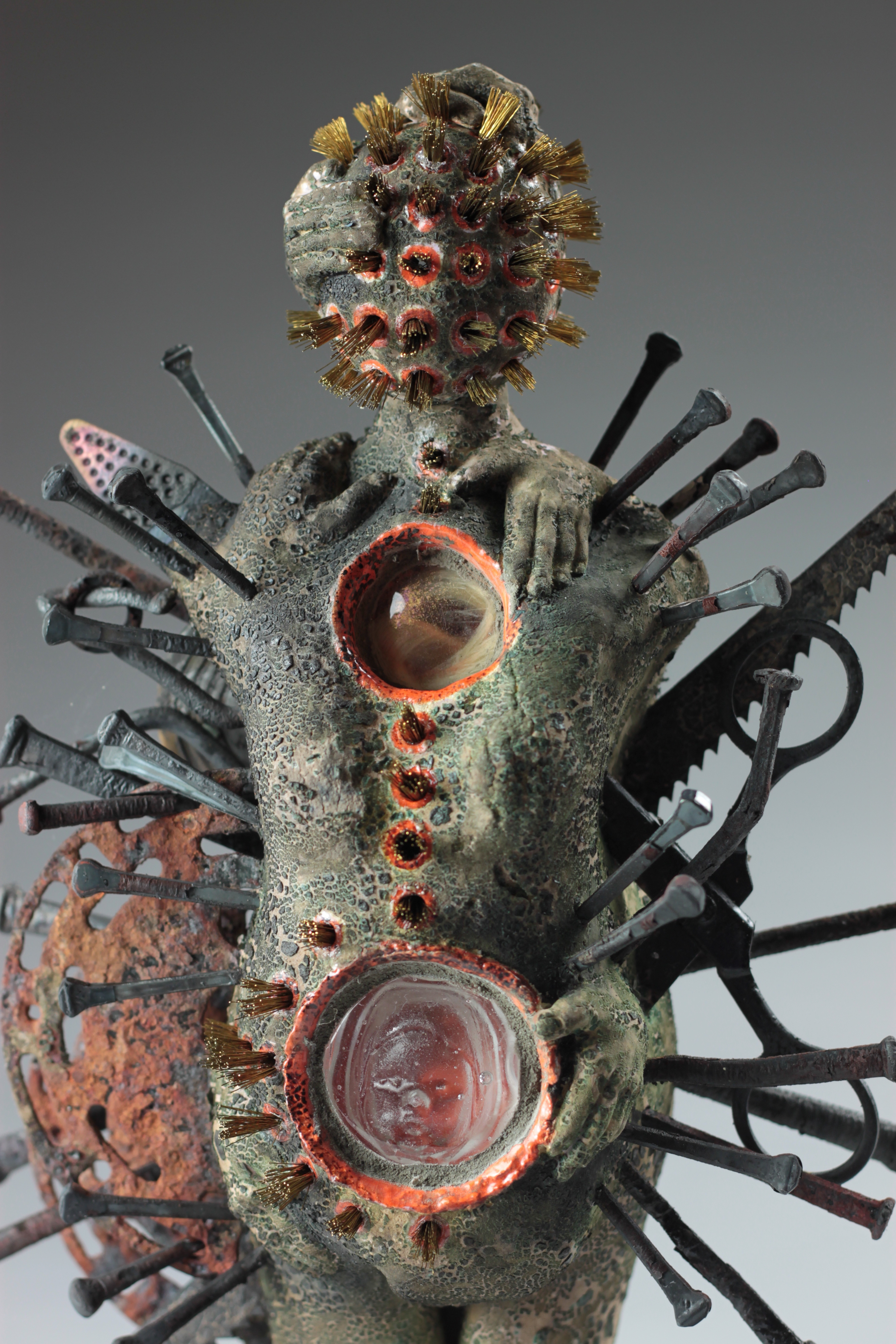 A mixed media sculpture, of a female form with gold metallic hair-like spiked on the head and metal spikes stuck in the body.