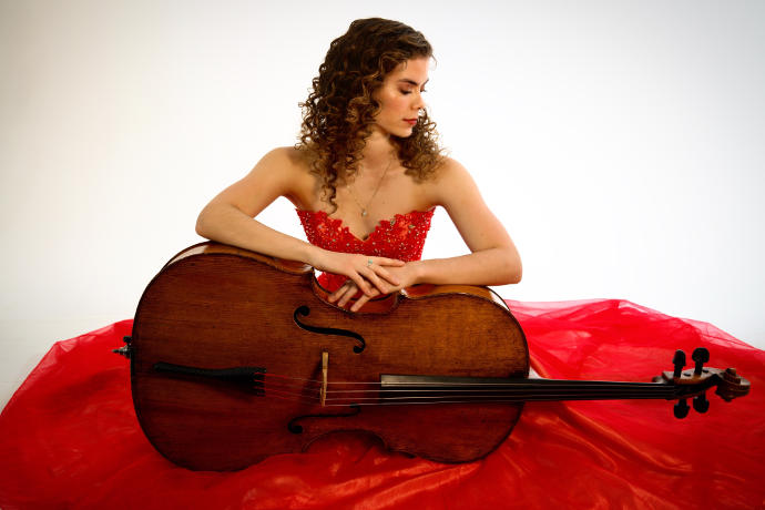 A photo of Katherine Audas, a young woman in a red dress holding a cello lengthwise across her lap