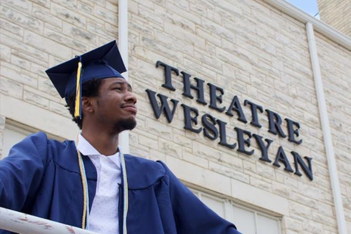A student in a cap and gown standing in front of the Theatre Wesleyan building