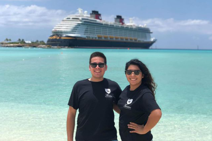 Two Texas Wesleyan alumni in TXWES shirts posing in front of a Disney cruise ship